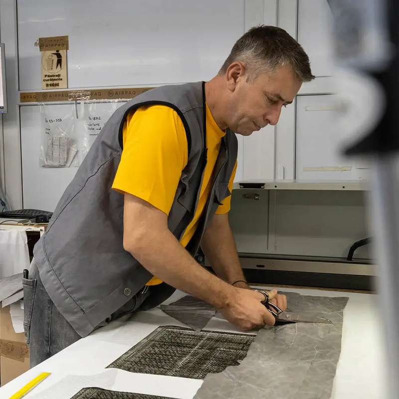 Person in gelbemShirt und grauer Weste schneidet mit einer Schere Airbagstoff auf einem Tisch in der Produktion.